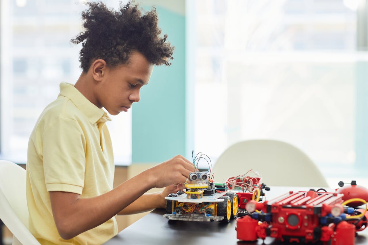 A young boy focusing on building a robotic project indoors, featuring toy cars and electronic components.